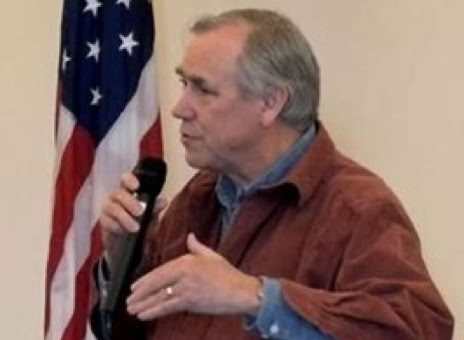 A person speaking into a microphone, standing next to an American flag.