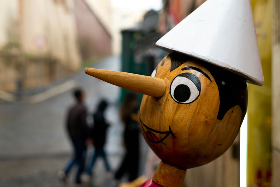 A wooden puppet with a long nose and a white hat is in the foreground, with blurred people walking in the background on a street.