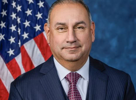 A man in a suit and tie stands in front of an American flag with a blue textured background.