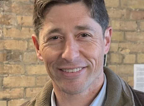A person with short brown hair, wearing a brown jacket over a light blue shirt, stands in front of a brick wall.