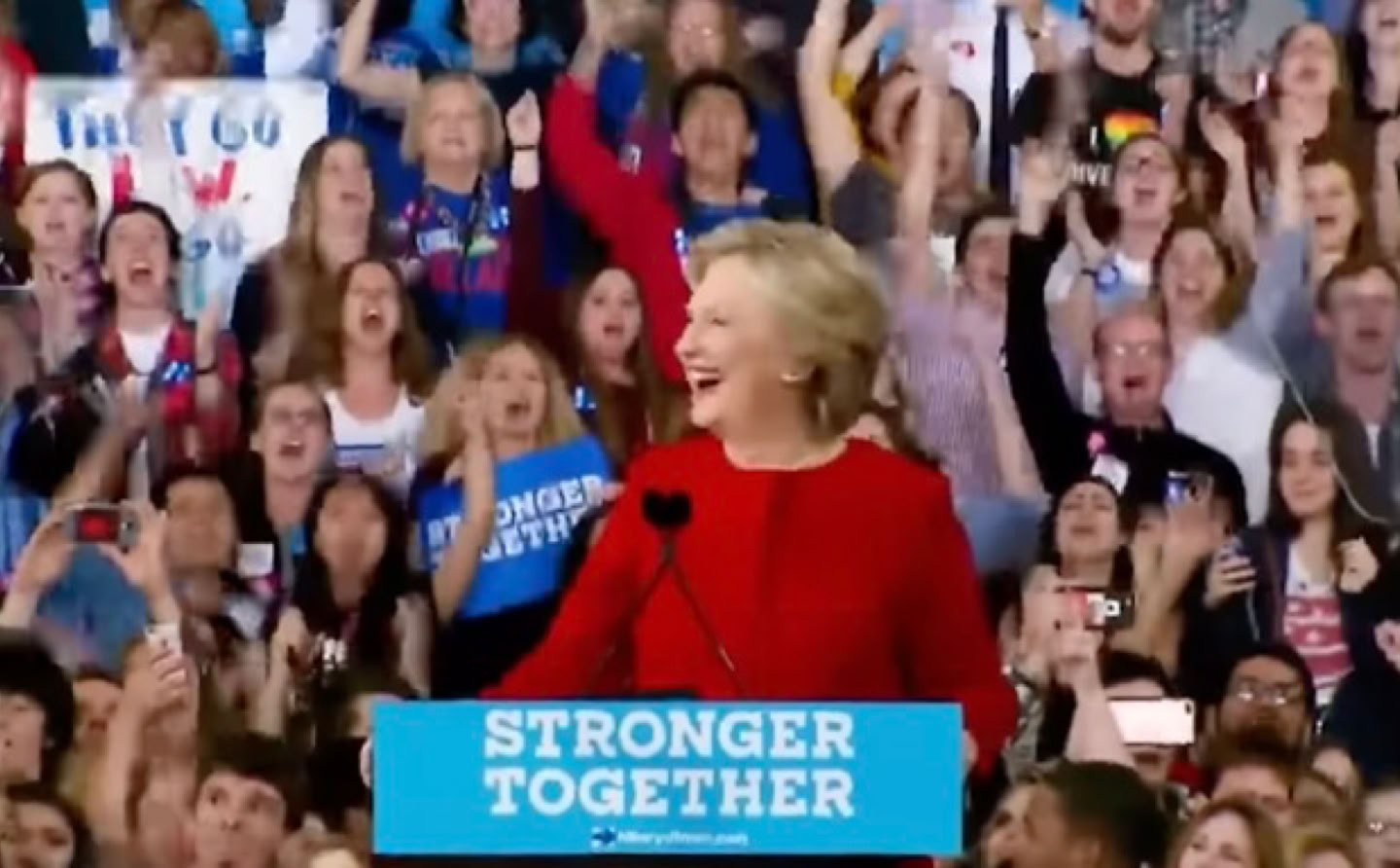 Hillary Clinton in a red outfit stands at a podium with a "Stronger Together" sign, surrounded by a cheering crowd holding signs.