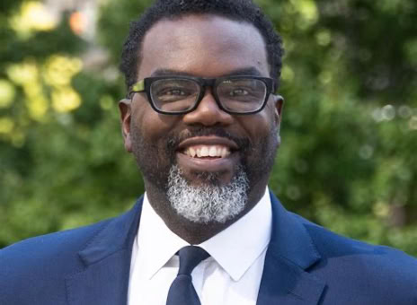A man with glasses and a beard, wearing a blue suit and tie, standing outdoors with greenery in the background.