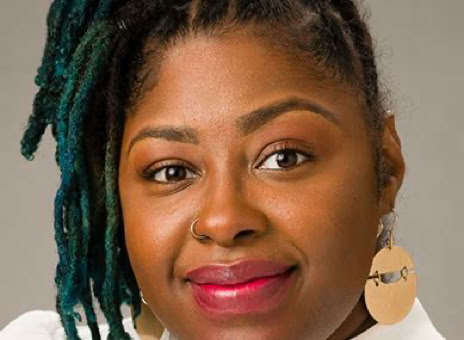A person with braided hair dyed in shades of blue and black, wearing a white top and large round earrings, against a neutral background.
