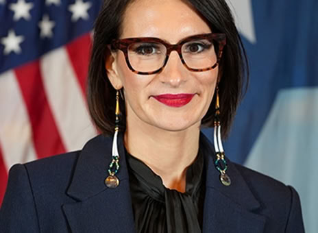 A person with short dark hair, wearing glasses, a dark suit, and a black blouse, standing in front of a U.S. flag and another flag with a white star.