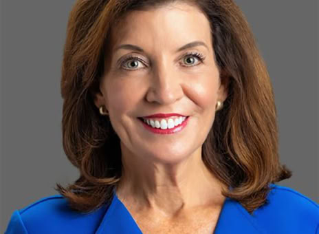 A woman with shoulder-length brown hair wearing a blue blazer, smiling against a gray background.