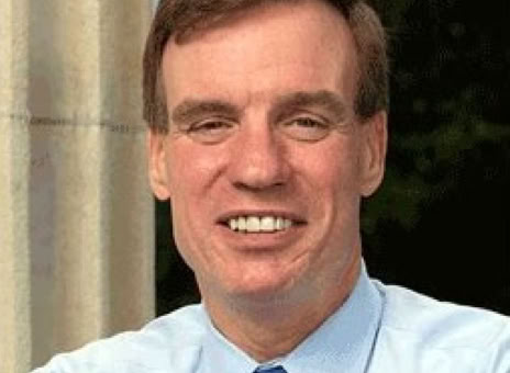 A man wearing a light blue shirt and blue tie, smiling, with a blurred background.