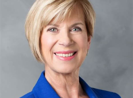 A person with short blonde hair wearing a blue blazer, smiling against a gray background.