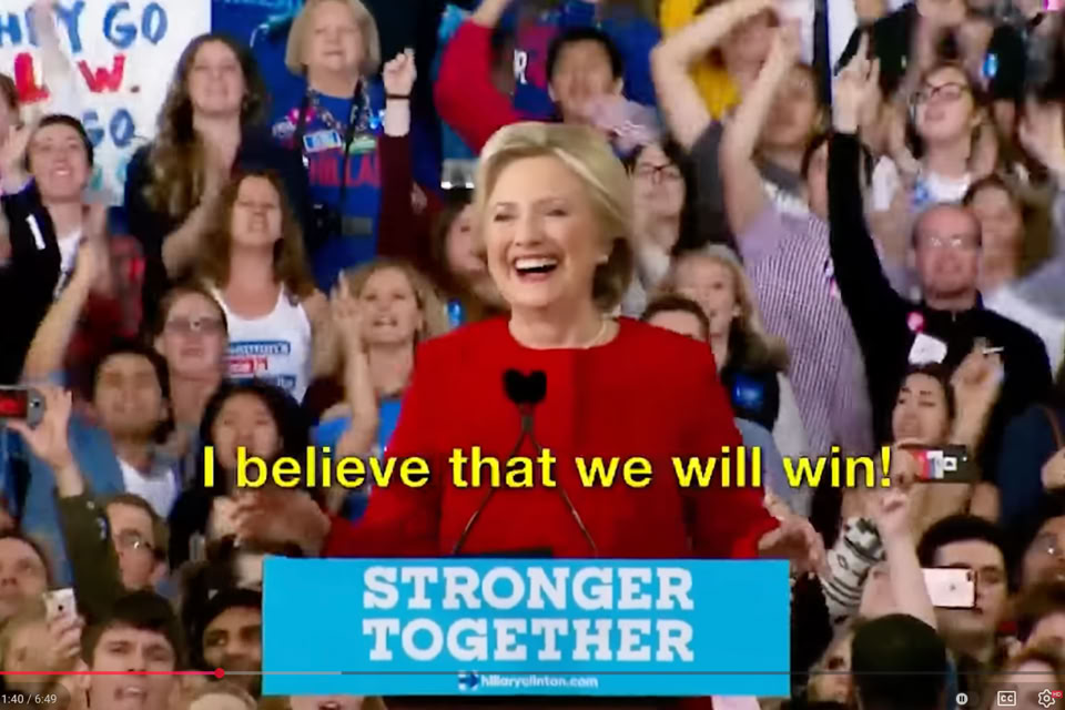 A person in a red outfit speaking at a podium with a "Stronger Together" sign, surrounded by a cheering crowd.