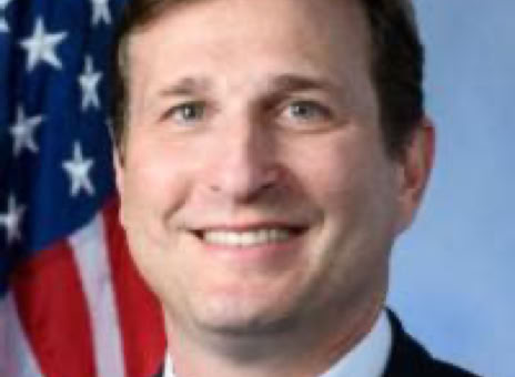 A man in a suit and tie smiling, with an American flag in the background.