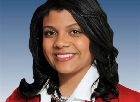 A person with long dark hair wearing a red blazer, white shirt, and a pearl necklace, smiling against a blue gradient background.
