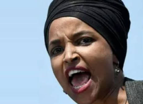 A person wearing a black headscarf is speaking with an open mouth against a clear blue sky.