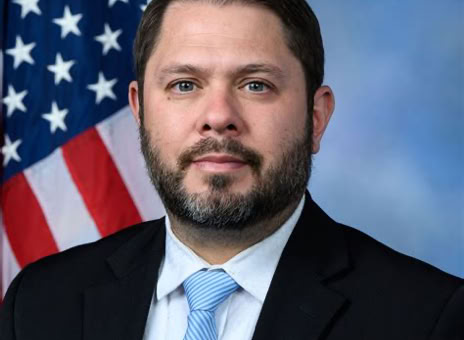 A man with a beard wearing a suit and light blue tie, standing in front of an American flag and a blue background.