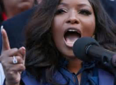 A woman speaking passionately into a microphone, wearing a blue outfit, with people in the background.