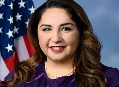 A woman with long brown hair wearing a purple top and a pearl necklace, standing in front of an American flag.